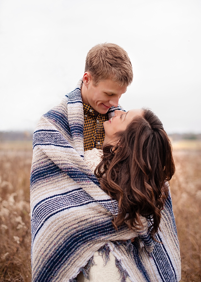 Engagement couple wrapped in blanket in field 