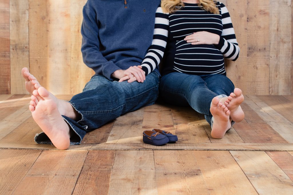 parents sitting holding hands on wood background with bare feet and baby shoes in front of them 