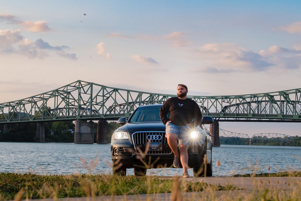highschool senior posing in front of his car by the river and a bridge