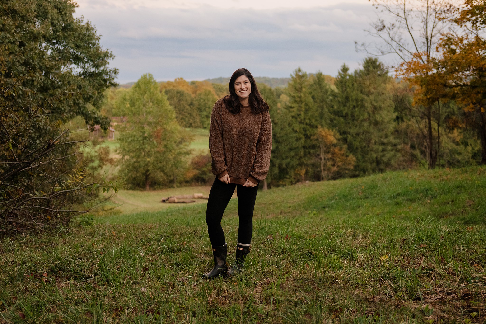 A woman in a brown sweater, smiling sweetly, stands in a field with beautiful scenery in the background.