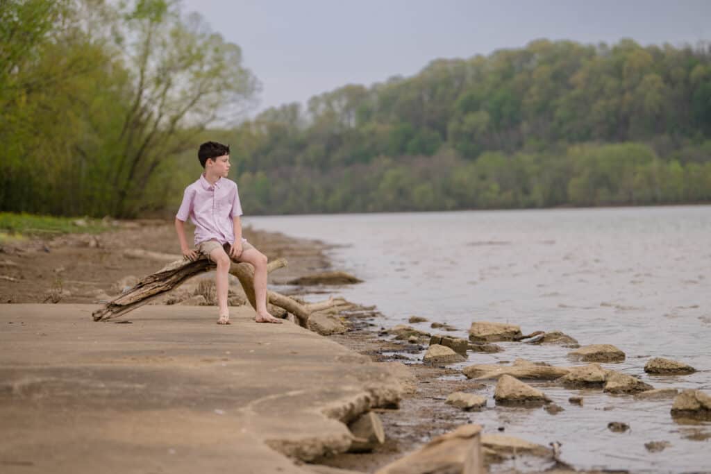 boy in pink shirt at riverbank sitting on driftwood