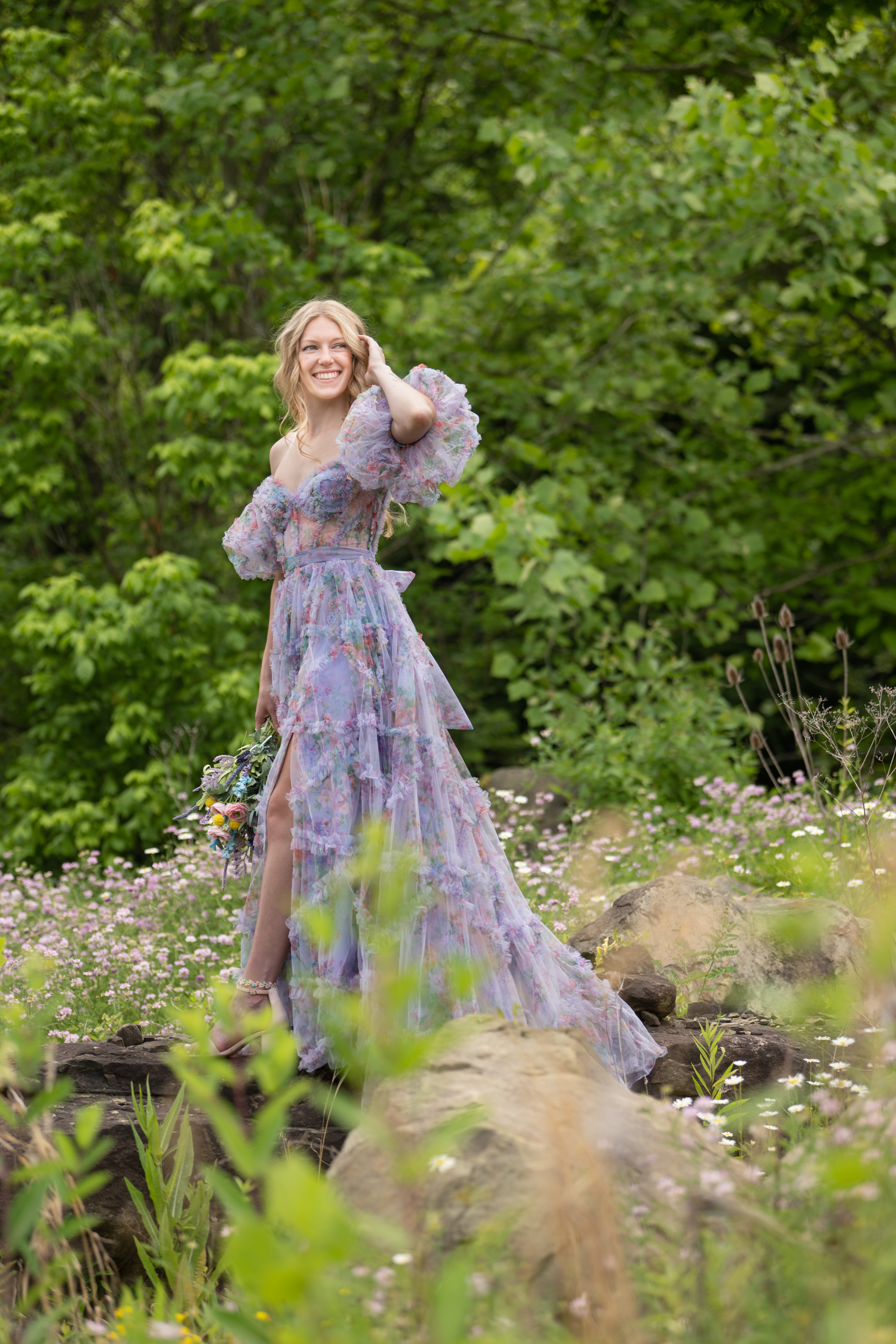 A fairy-like lady in a mesh flowery purple gown smiles as she stands on top of a log surrounded by beautiful flowers and trees.