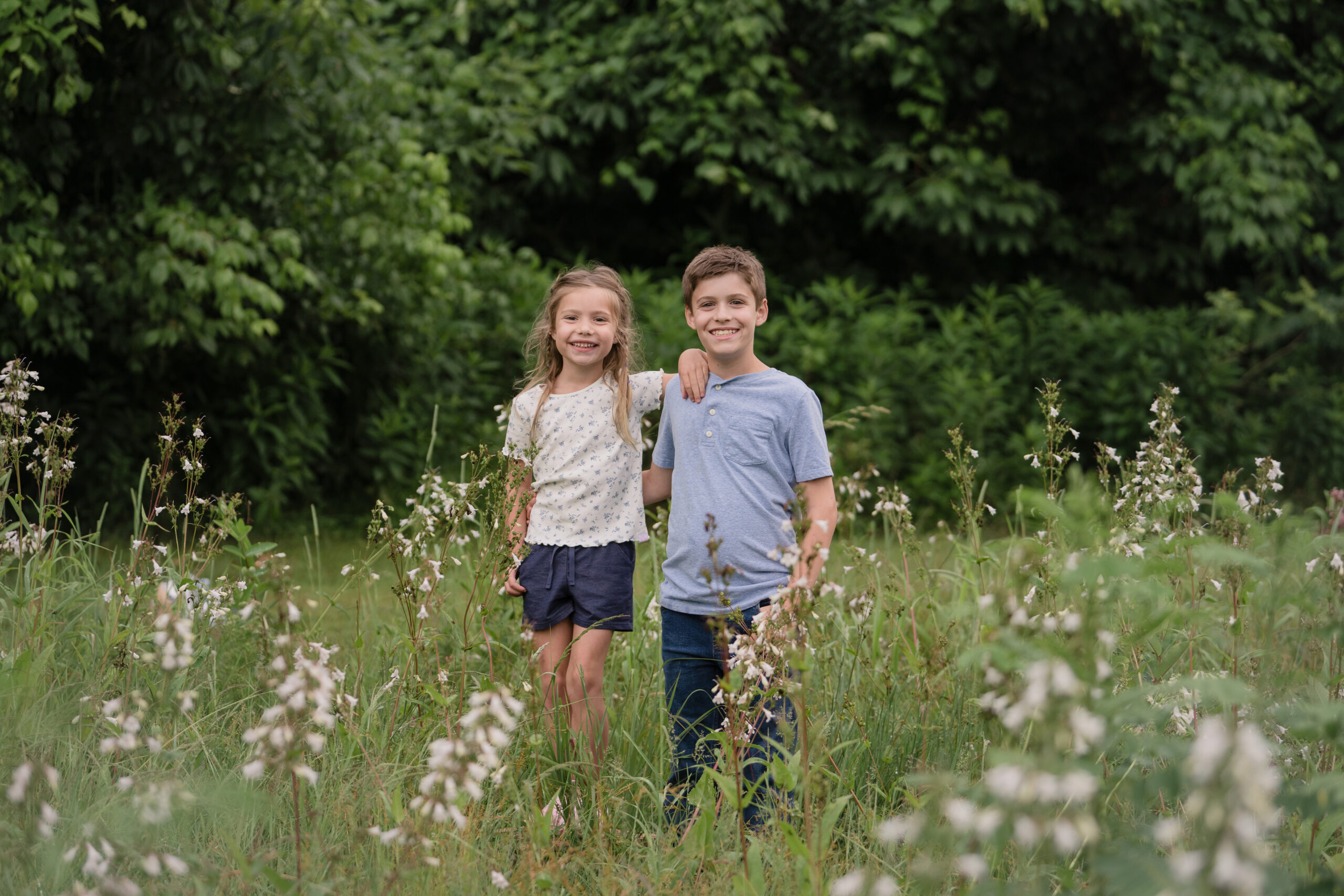 girl standing in the flower field with her arms placed on the boy's shoulder aknd them smiling in front. Boy posing in a field for a photography session near Marietta, OH. Lori Pickens Photography took the picture and is based out of Parkersburg, WV