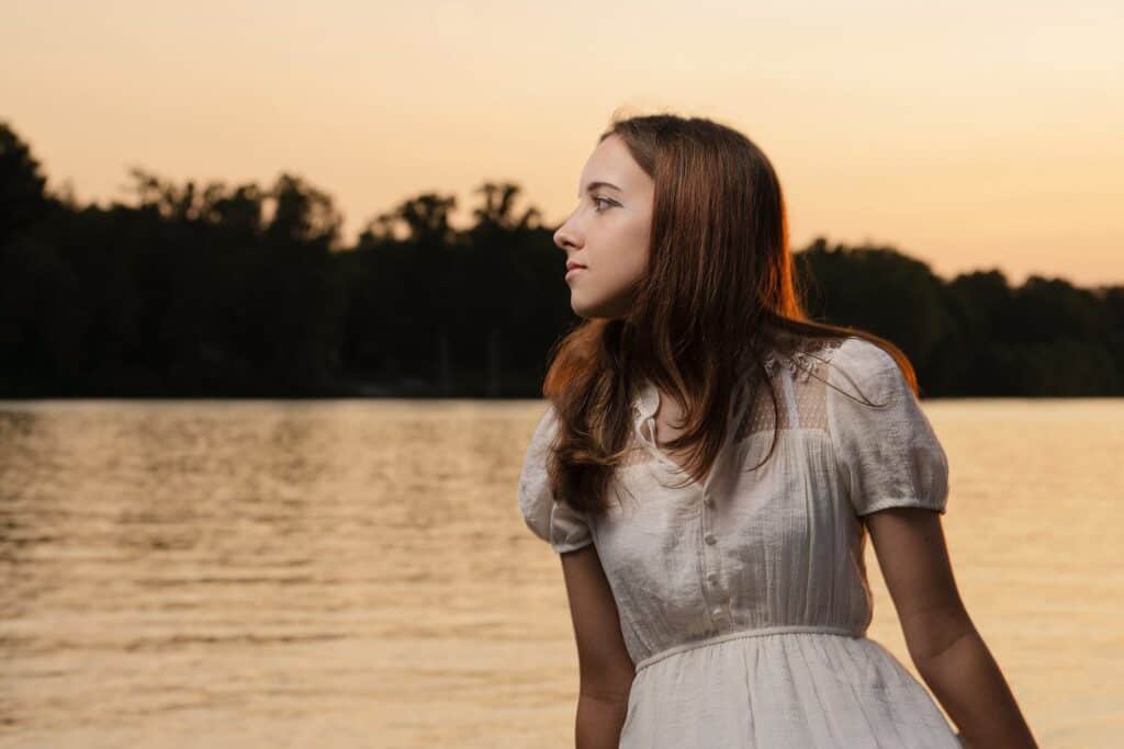 Girl looking out over the waterfront near Marietta, OH in her Senior Photography Session with Lori Pickens Photography.  She is wearing a white dress and has brown hair.  She has the evening sun creating beautiful light