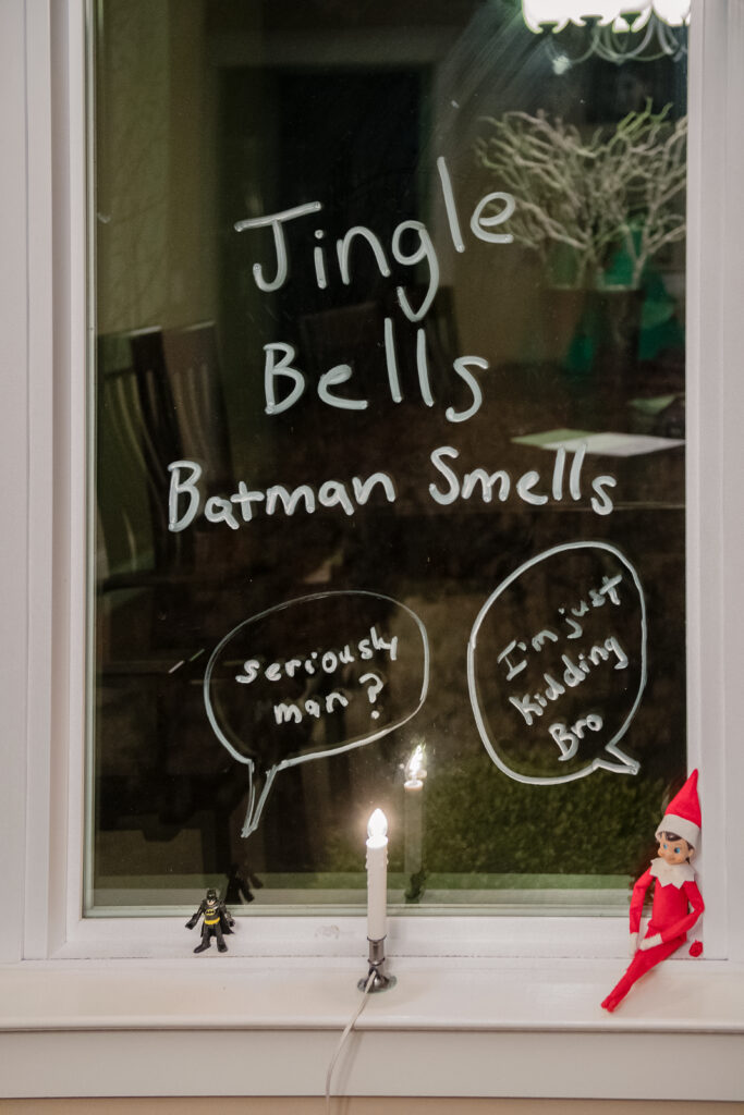 Elf on the Shelf and little batman. Window reads Jingle Bells, Batman Smells, Batman says seriously man, and elf on the shelf says I'm just kidding bro
