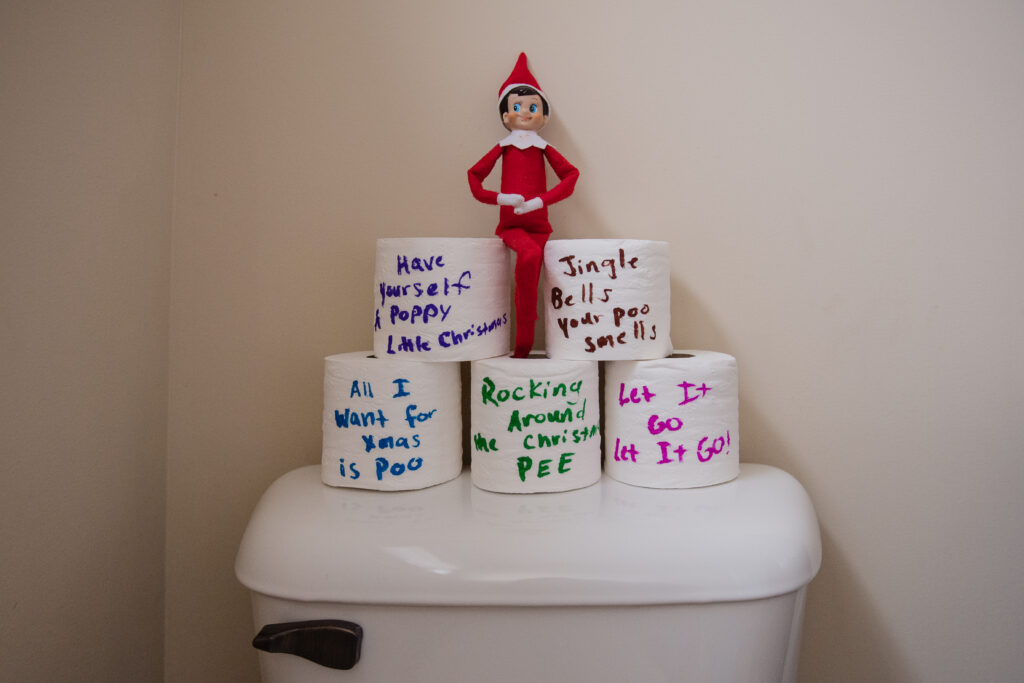 Elf on the shelf sitting on a tower of toilet paper.  It has written jokes on them.