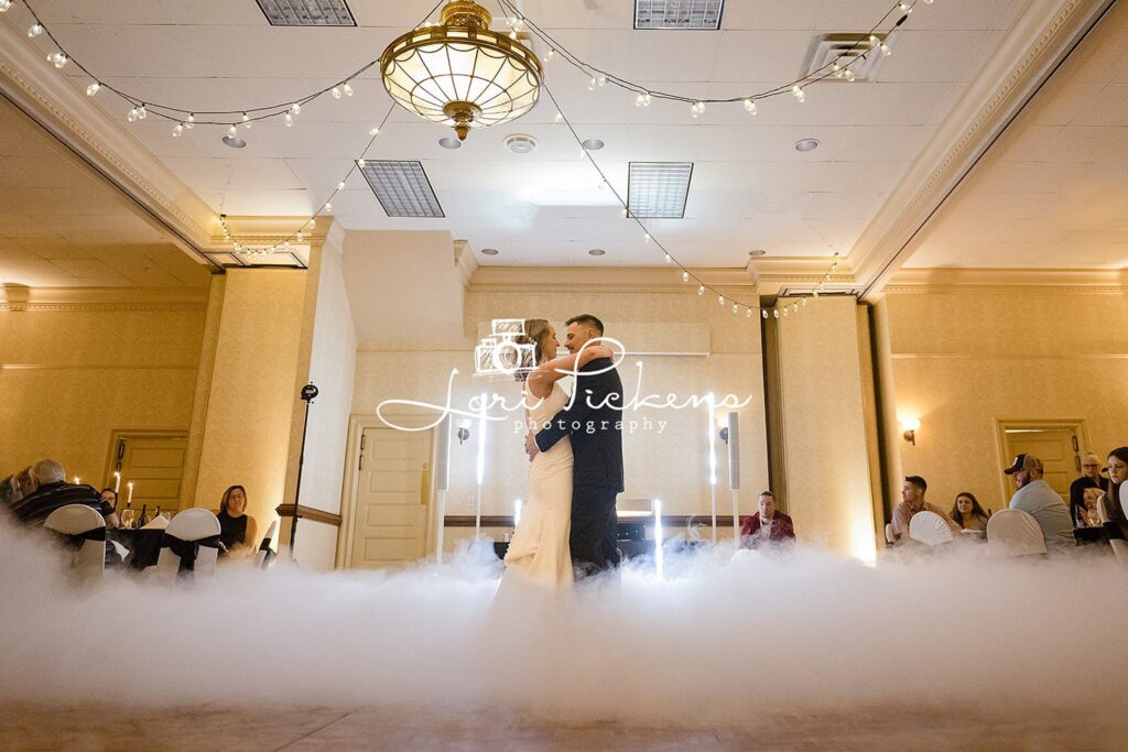 Newlyweds dance in a ballroom covered in ground fog surrounded by guests