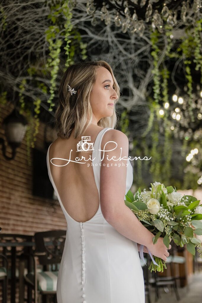 A bride in a white silk dress stands under a patio holding her bouquet and looking over her shoulder