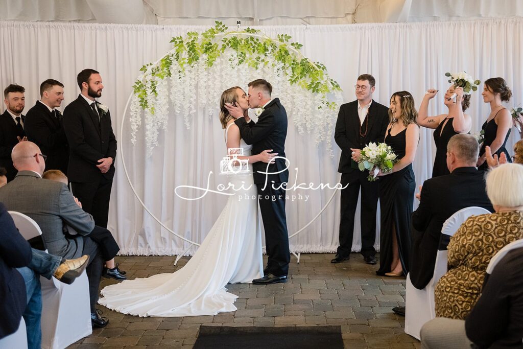 Newlyweds kiss in front of a simple white floral arch to end their wedding ceremony