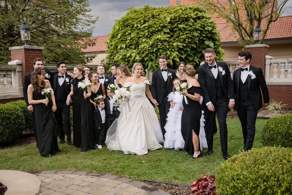 A wedding party in black suits and dresses walks through a garden chatting and laughing at a Blennerhassett Hotel wedding