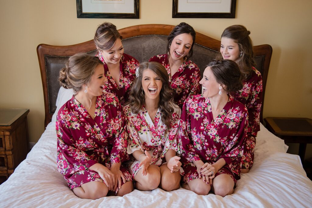 A bride in a pink floral robe kneels on a bed laughing with her bridesmaids in matching robes at a Blennerhassett Hotel wedding