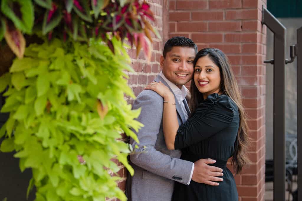 Newly engaged couple hugs while leaning against a brick wall behind hanging plants