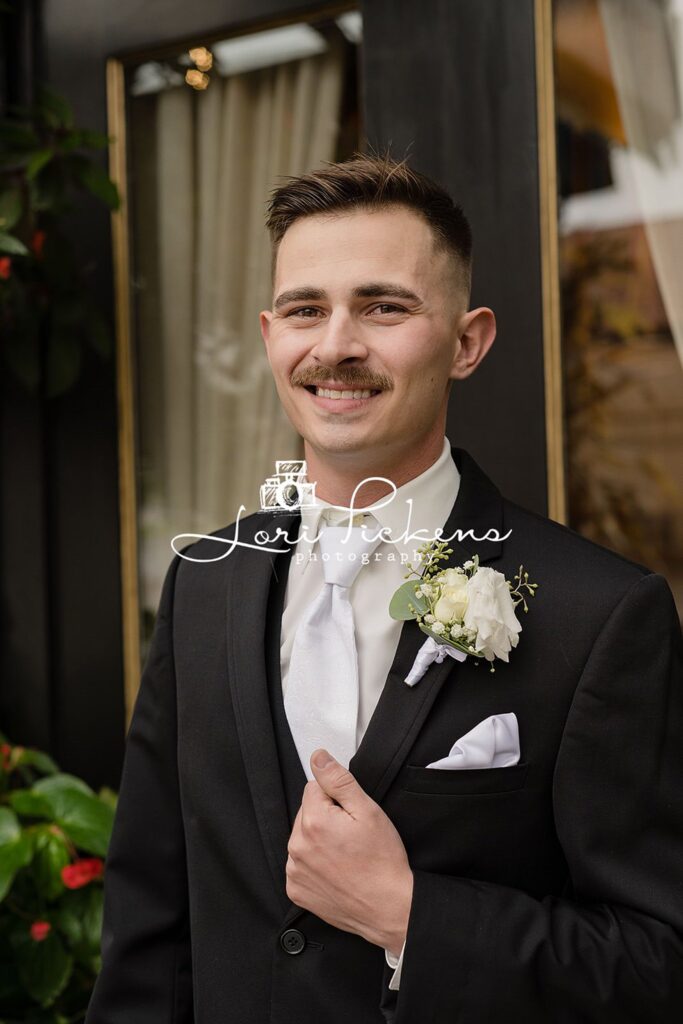 A groom in a black suit and white tie stands by a window holding his lapel at one of the wedding venues west virginia
