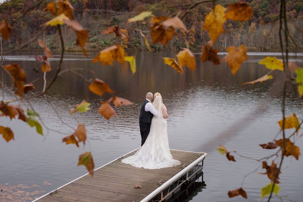 Bride and groom stand embraced on edge of boat dock by water surrounded by fall leaves near wedding venues in Charleston WV