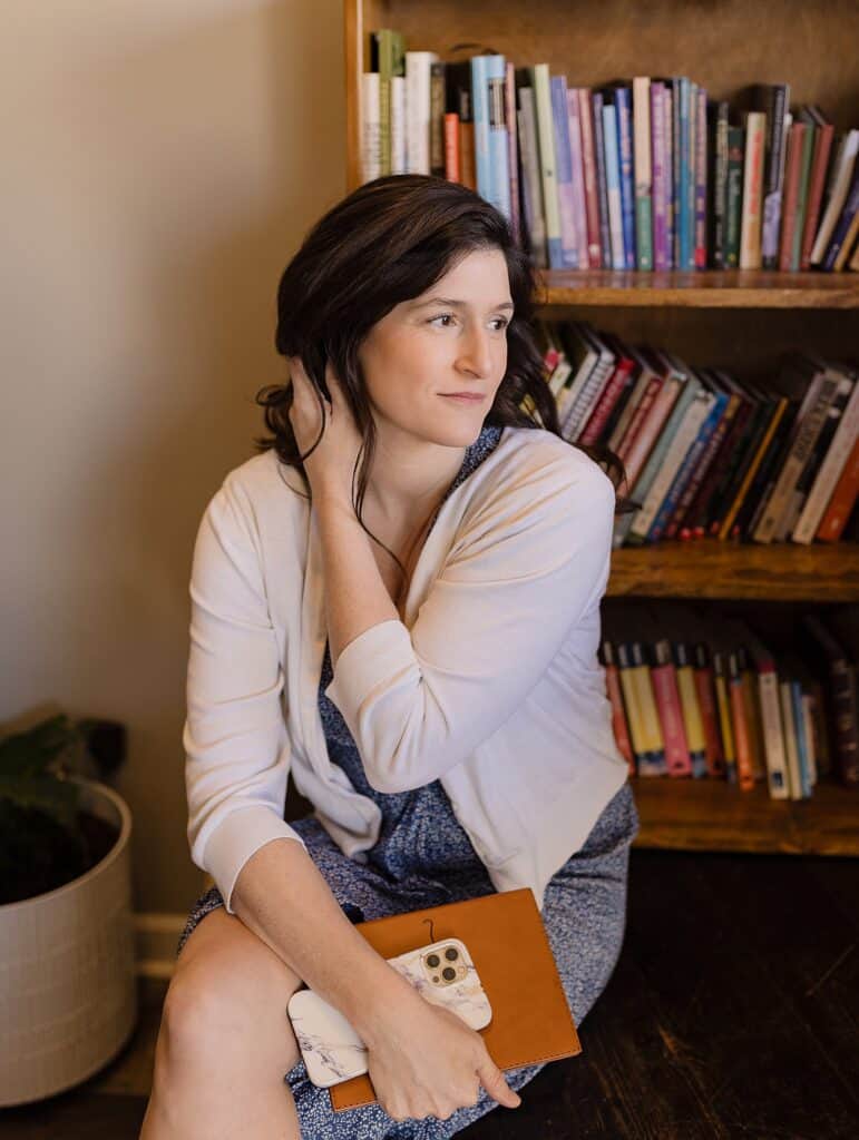 Lori Pickens - photographer and podcast host - sitting on wood step in front of a book case holding a phone and notebook in her had. 