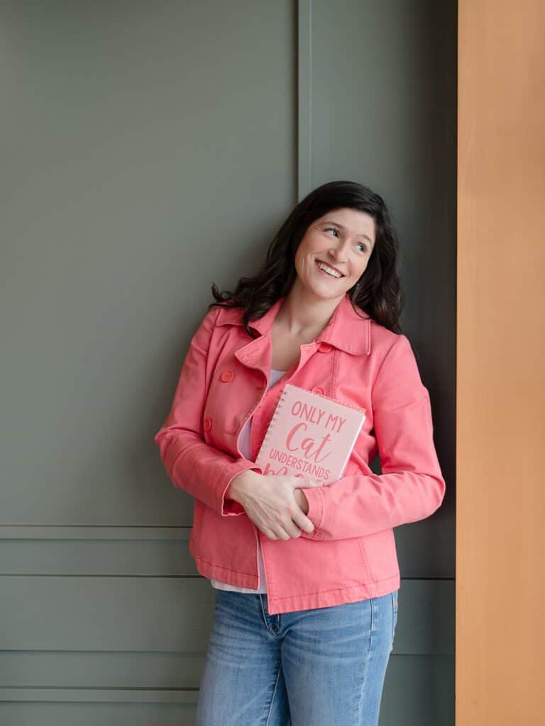 Woman in bright pink coral jacket with dark brown hair leaning on a teal green wall holding a notebook that reads only my cat understands me while looking out the window on the side related to the podcast episode about common branding mistakes photographers often make