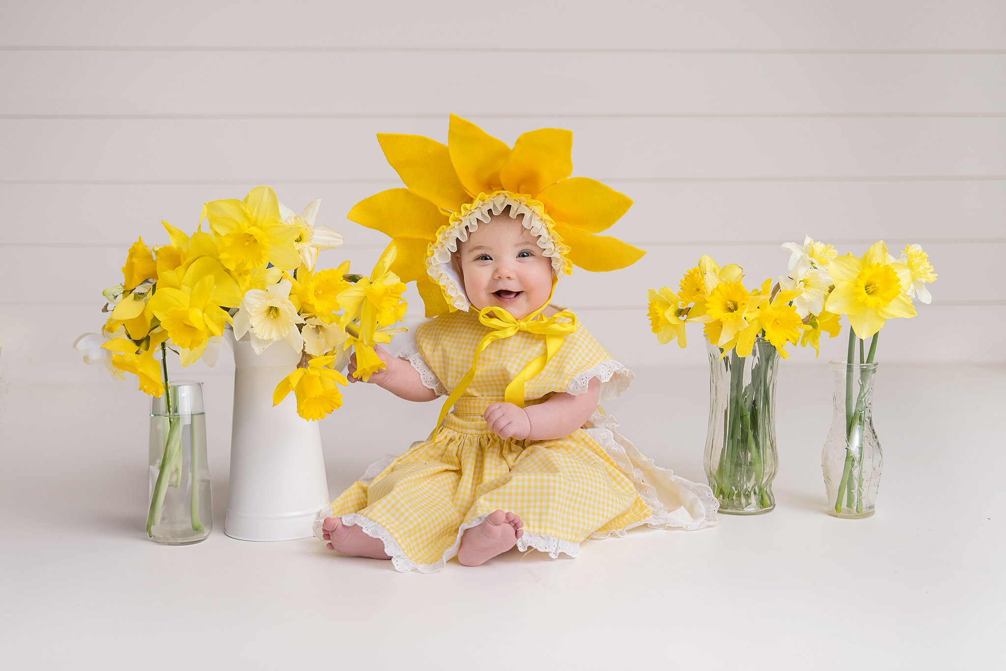 sitting baby in yellow dress and hat on white background with vases and buckets of daffodils beside her