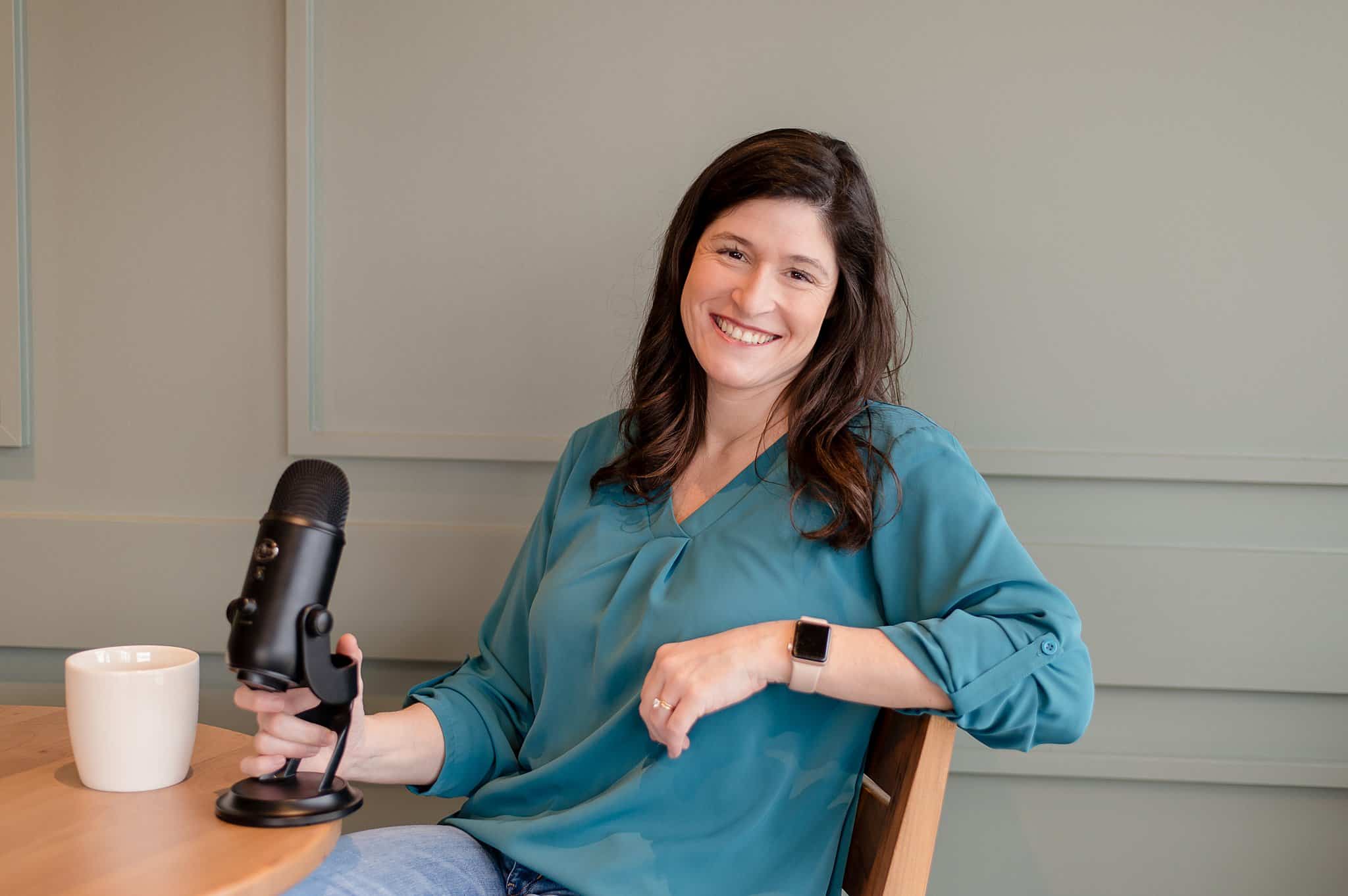 Lori Pickens, woman in a green top sitting in a chair smiling with a podcast microphone, and a mug of coffee