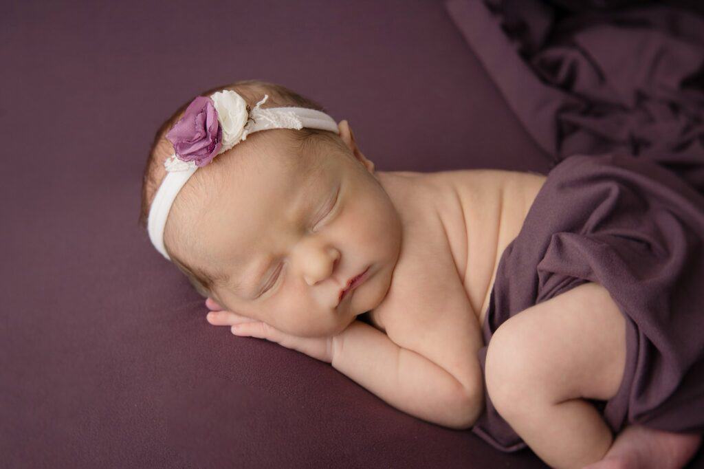 baby in a floral headband on a plum colored pillow with a sheet draped over the legs.