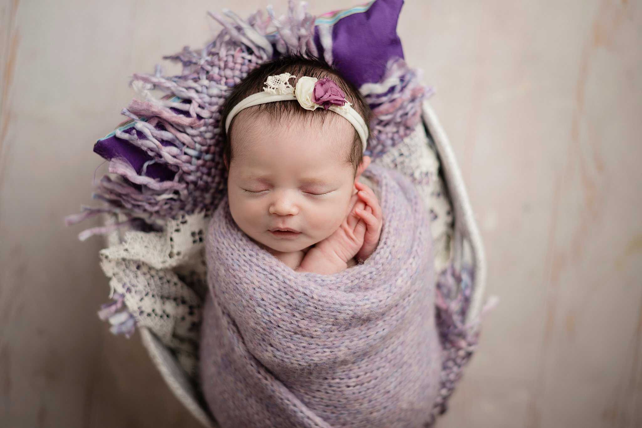 Baby wrapped in a purple swaddle in a basket with floral headband