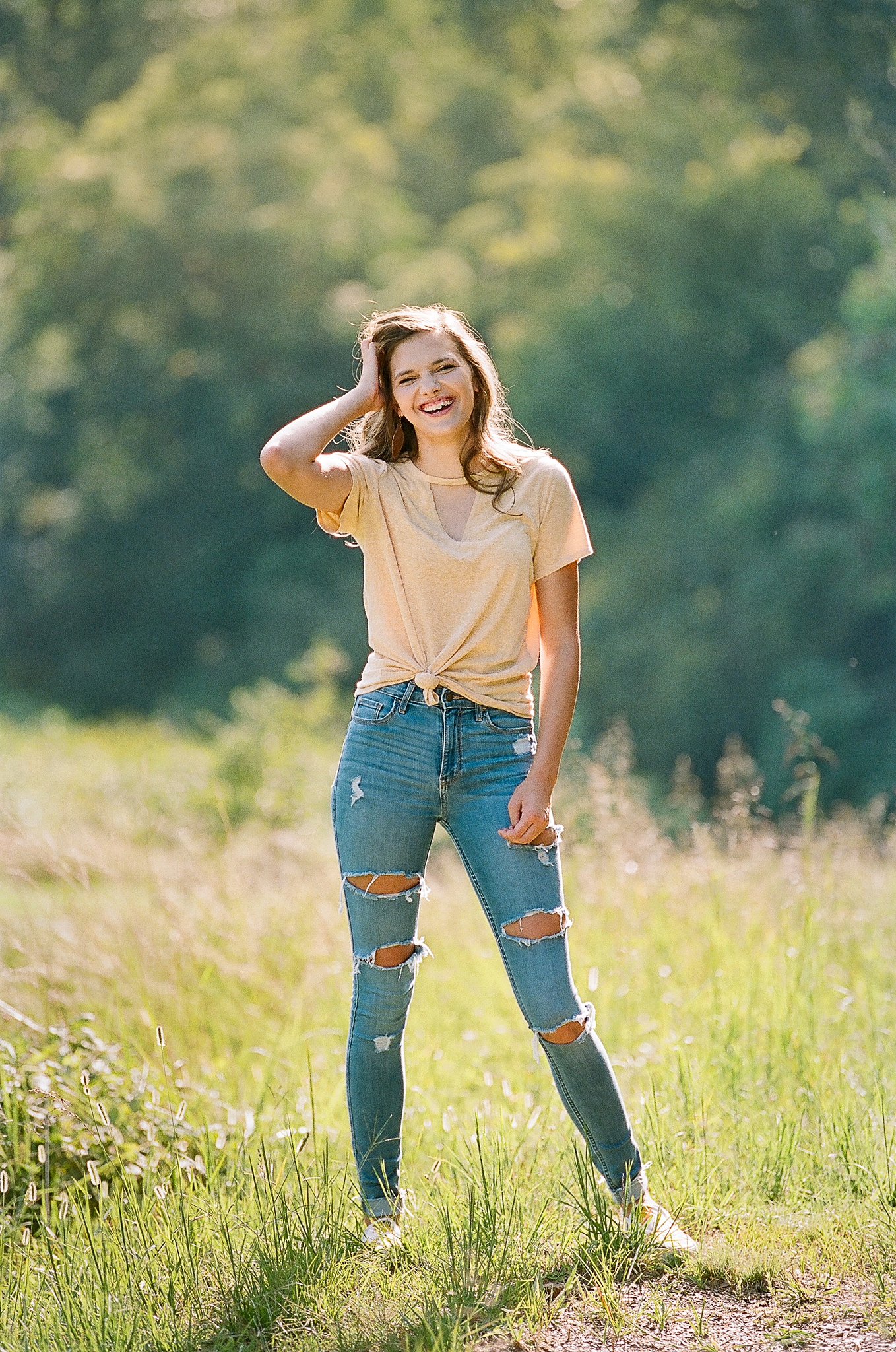A happy high school senior laughs while standing in a meadow with a hand in her hair after some tanning in parkersburg wv