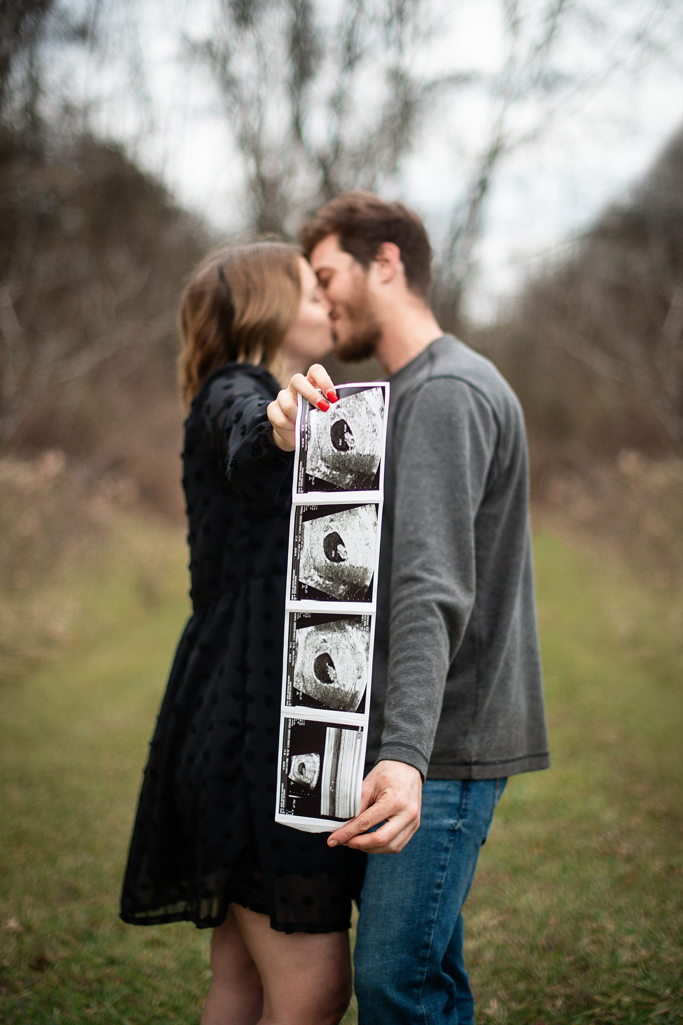 A happy pregnant couple kiss while holding out their strip of sonogram images in a forest