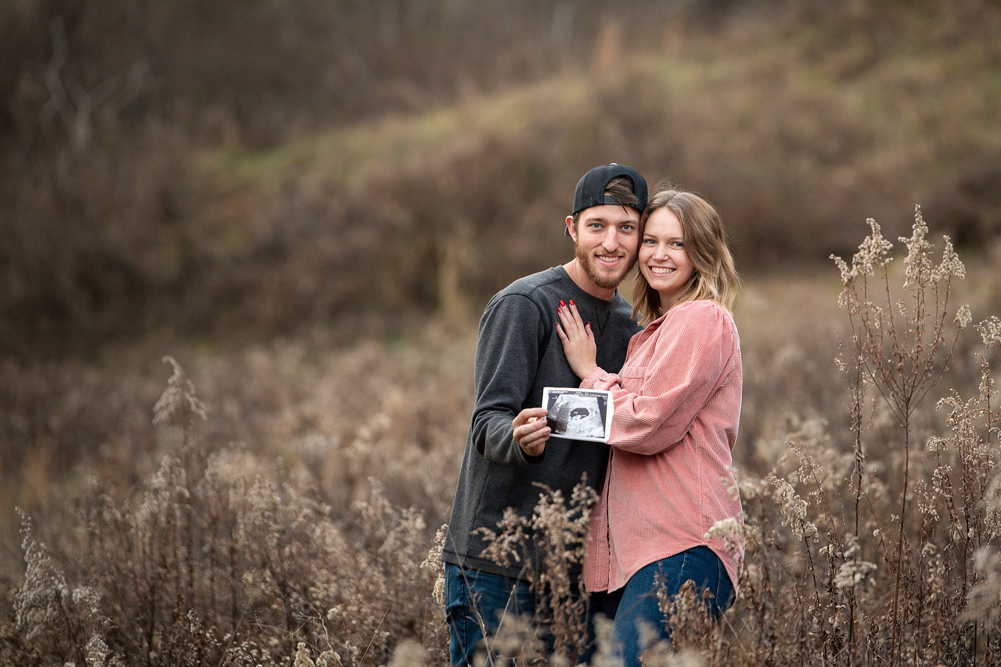 A happy pregnant couple stand in a meadow of tall grasses in fall holding out their sonogram after getting a 4d ultrasound in athens ohio