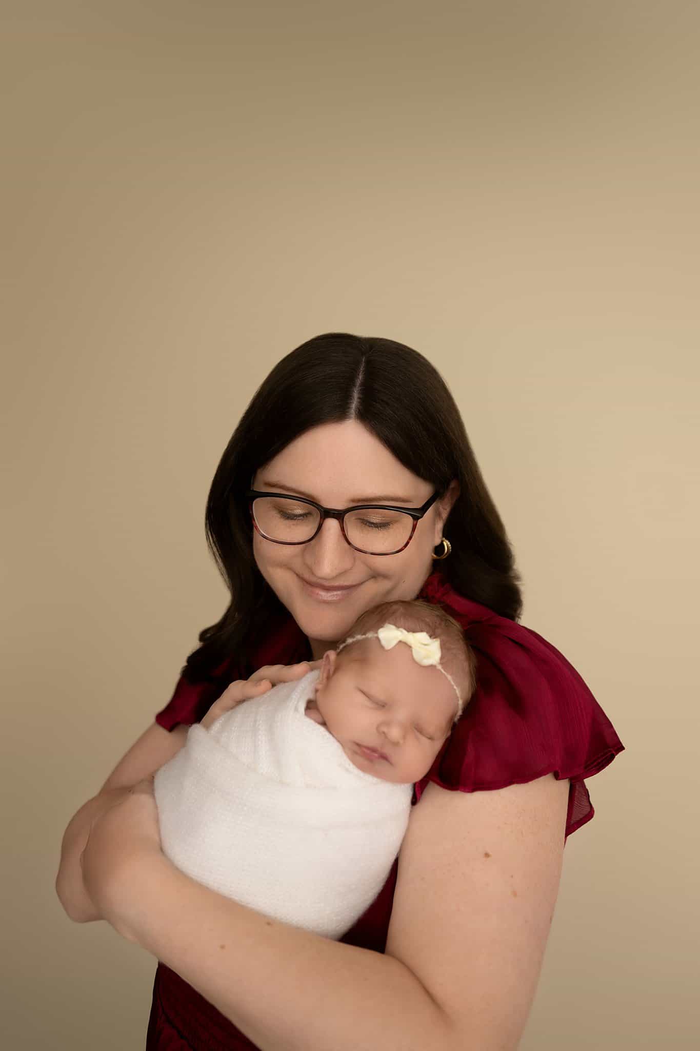 A happy new mom cradles her sleeping newborn under her chin on her shoulder in a white swaddle and bow headband