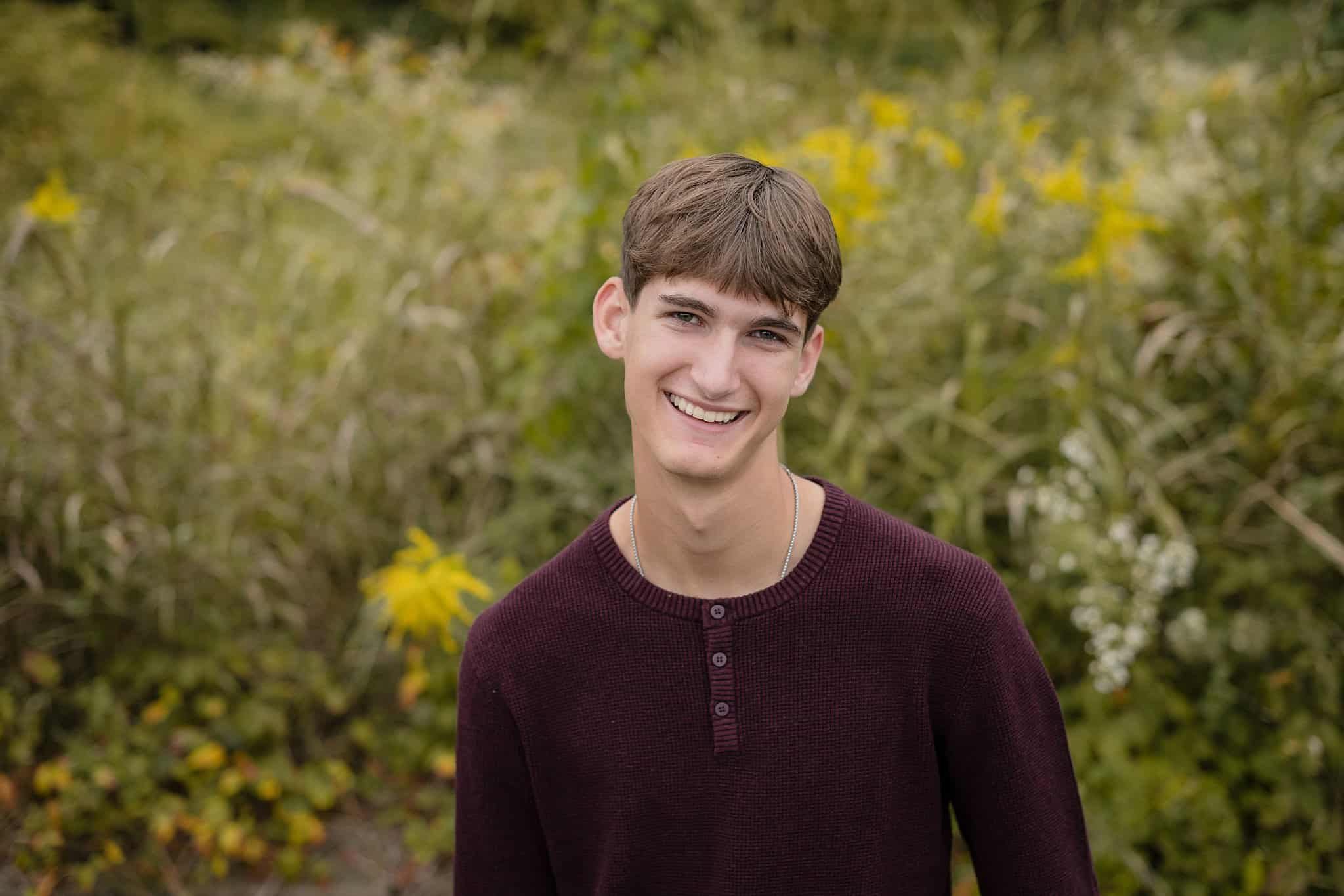 A happy high school senior smiles in a purple henley on the edge of tall grasses and wildflowers