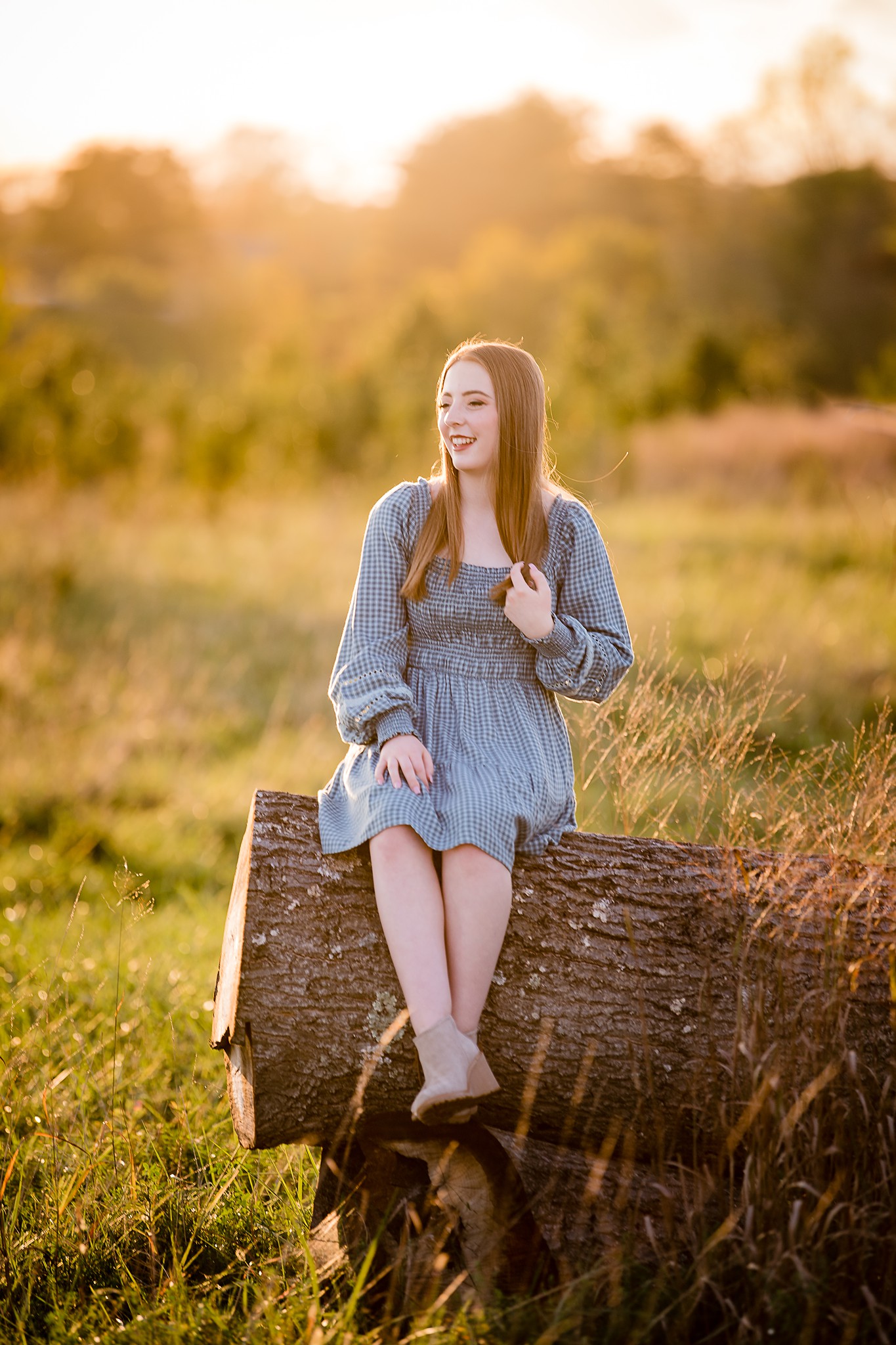 A happy teenager in a blue dress sits on a fallen log at sunset in a meadow
