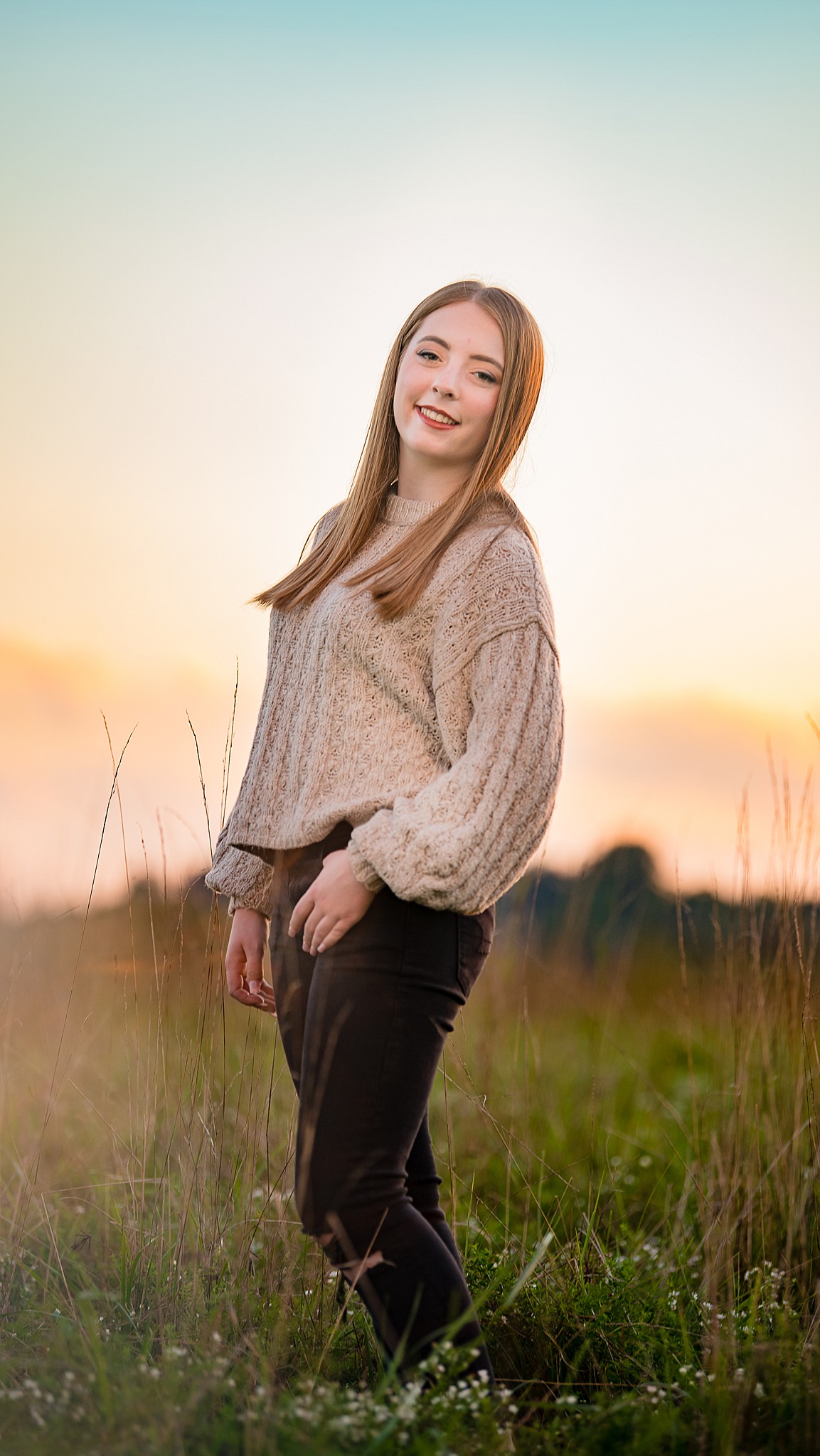 A high school senior in a tan sweater and black pants smiles while walking a field at sunset after visiting boutiques in parkersburg wv