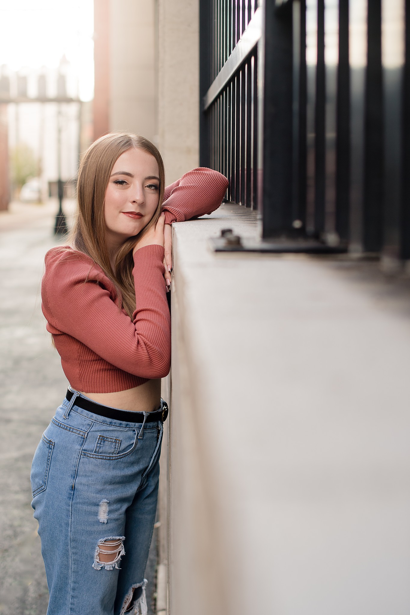 A high school senior in a pink sweater and jeans leans against a railing at head height after visiting boutiques in parkersburg wv