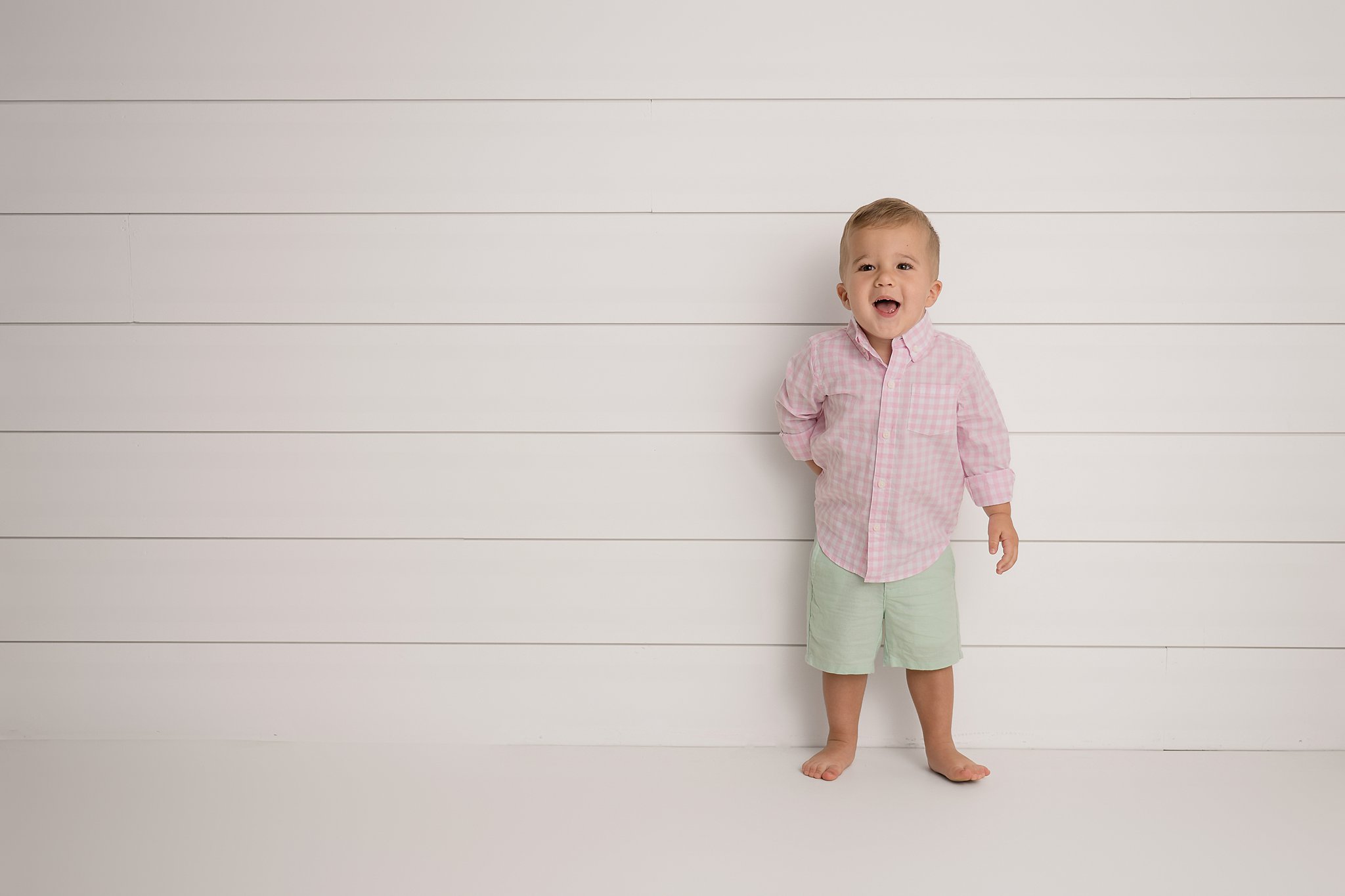 A toddler boy in green shorts and pink shirt leans against a wall barefoot with a hand behind his back