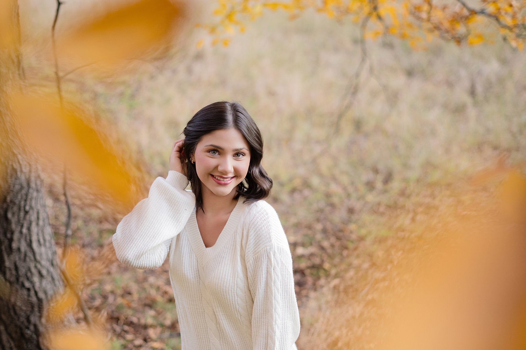 A smiling high school grad in white pulls her hair behind her ear in a forest in fall