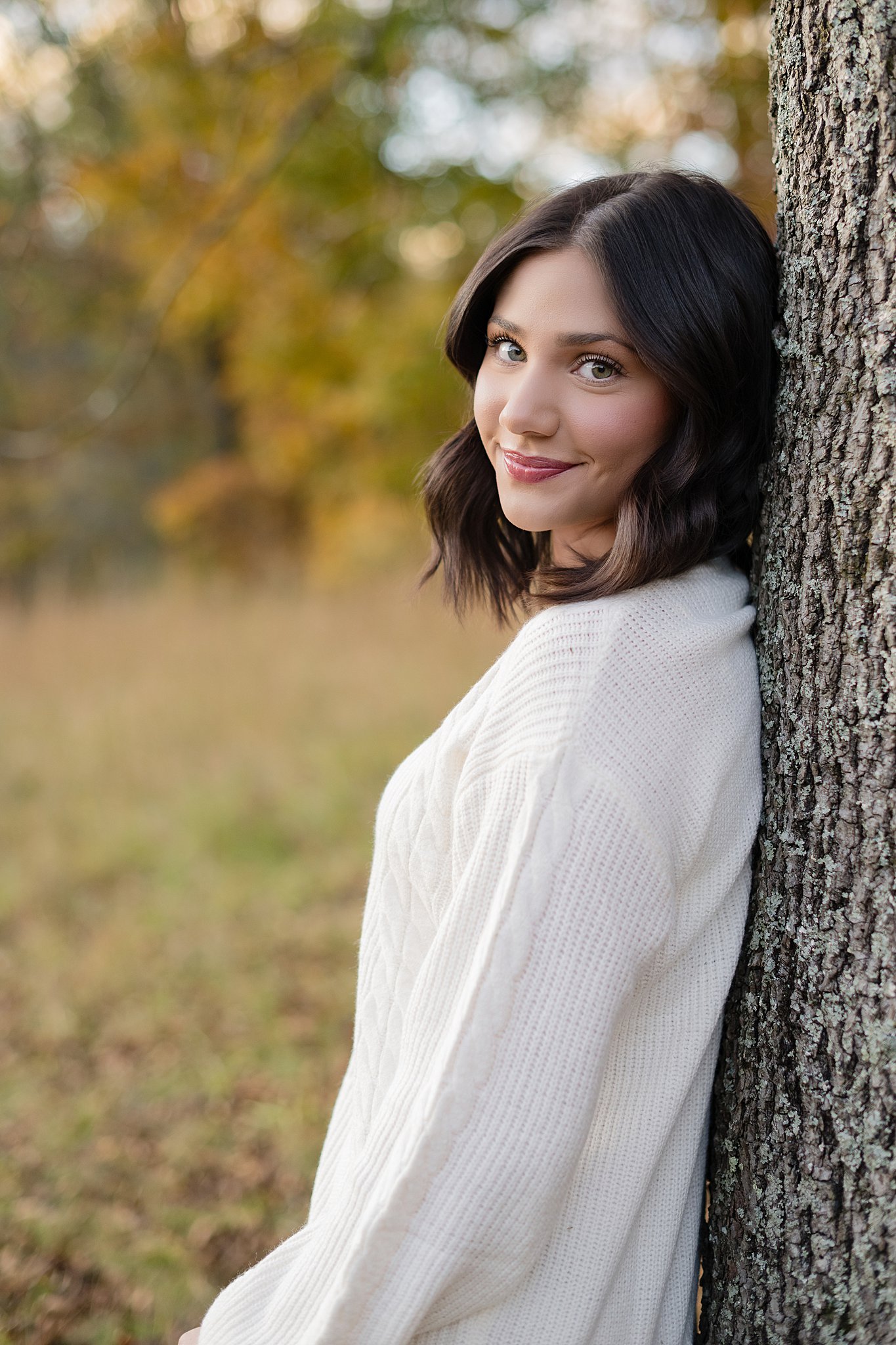 A happy high school senior leans against a tree smiling over her shoulder in a white dress after visiting graduation party venues in Parkersburg, WV