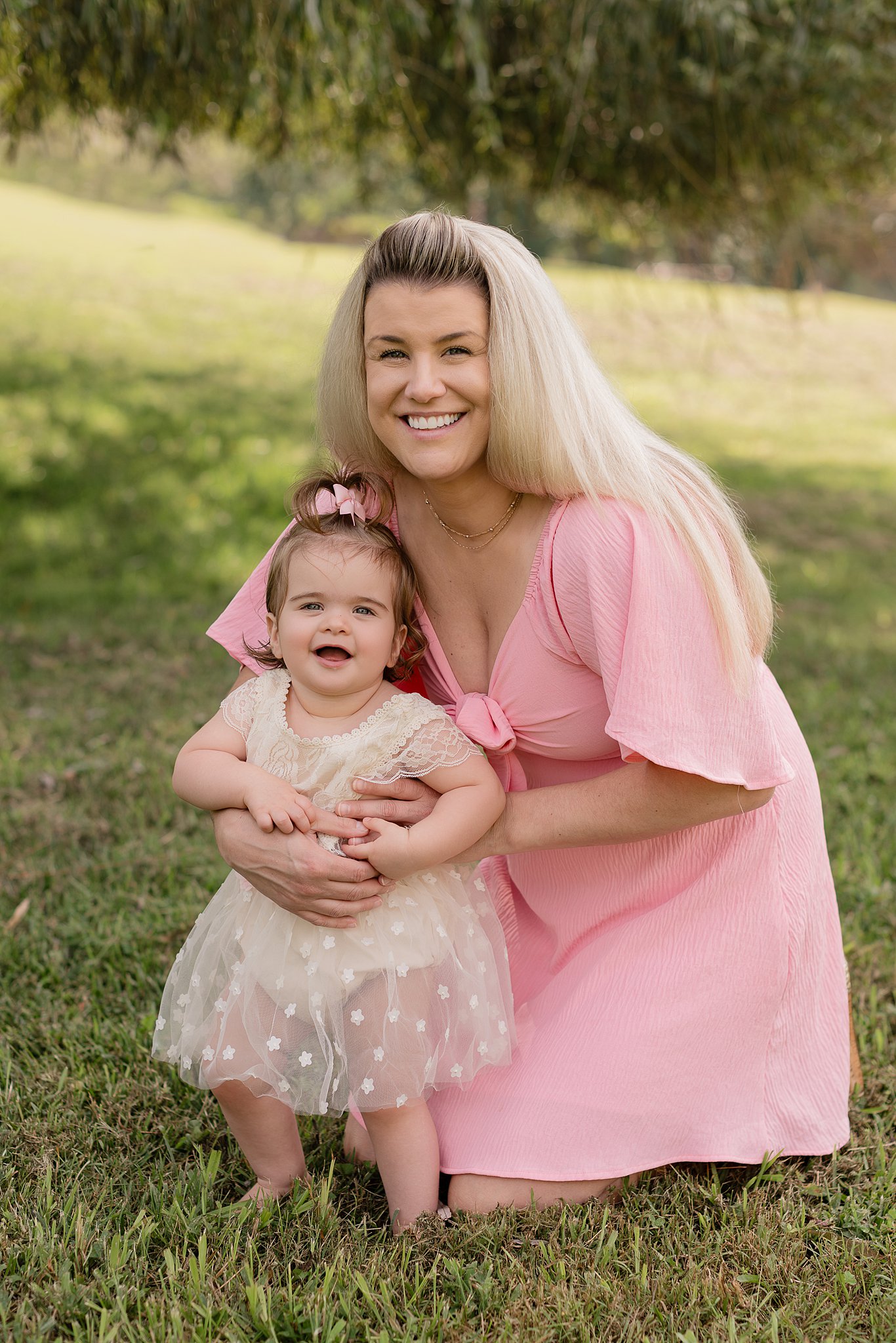 A happy mom in a pink dress hugs her smiling infant in a lawn in a white star covered gown before visiting Kid friendly restaurants in parkersburg, wv