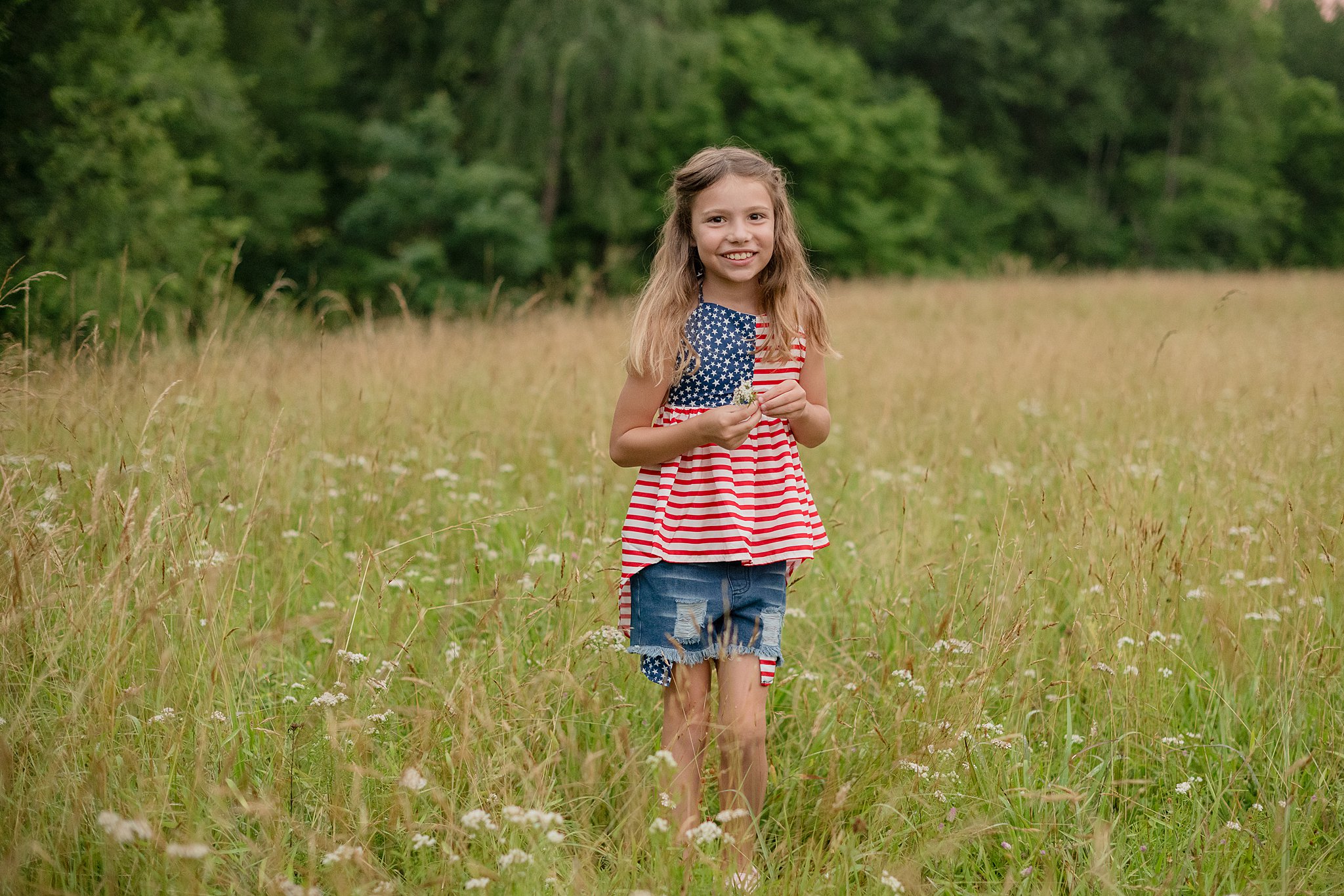 A young girl in american flag shirt picks wildflowers in a meadow at sunset after visiting a montessori school in parkersburg, wv