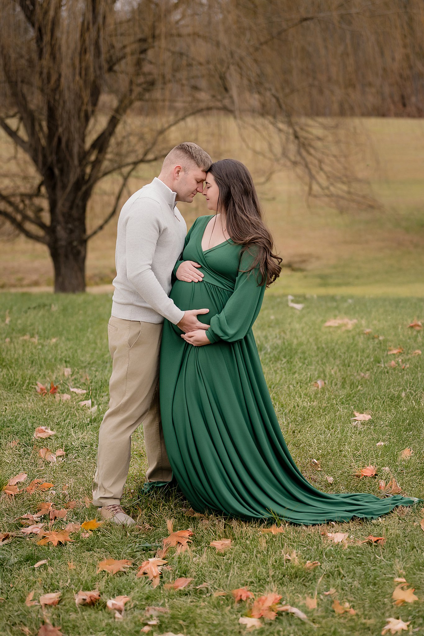 Happy expecting parents snuggle touching foreheads and holding the bump in a green maternity gown on a hill in a park after finding OBGYNs in Parkersburg, WV