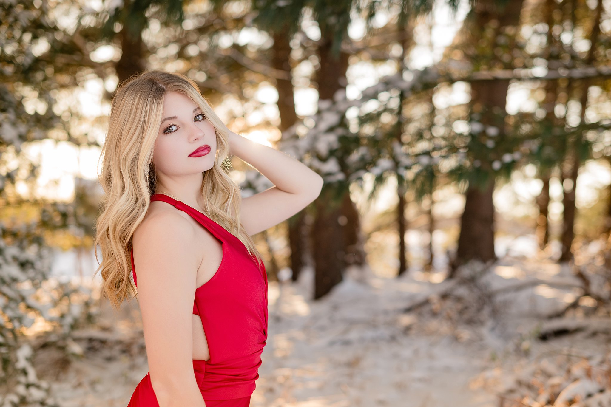 A woman runs fingers through her hair while standing in a red gown in the snow at sunset