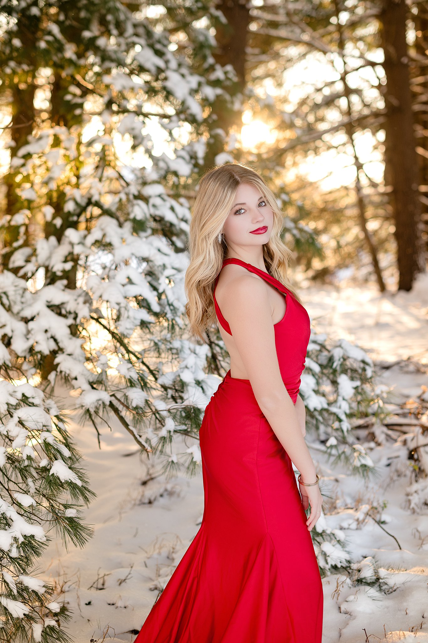 A high school senior in a red gown stands in a snow covered forest at sunset smiling over her shoulder after finding hair salons in Belpre, OH