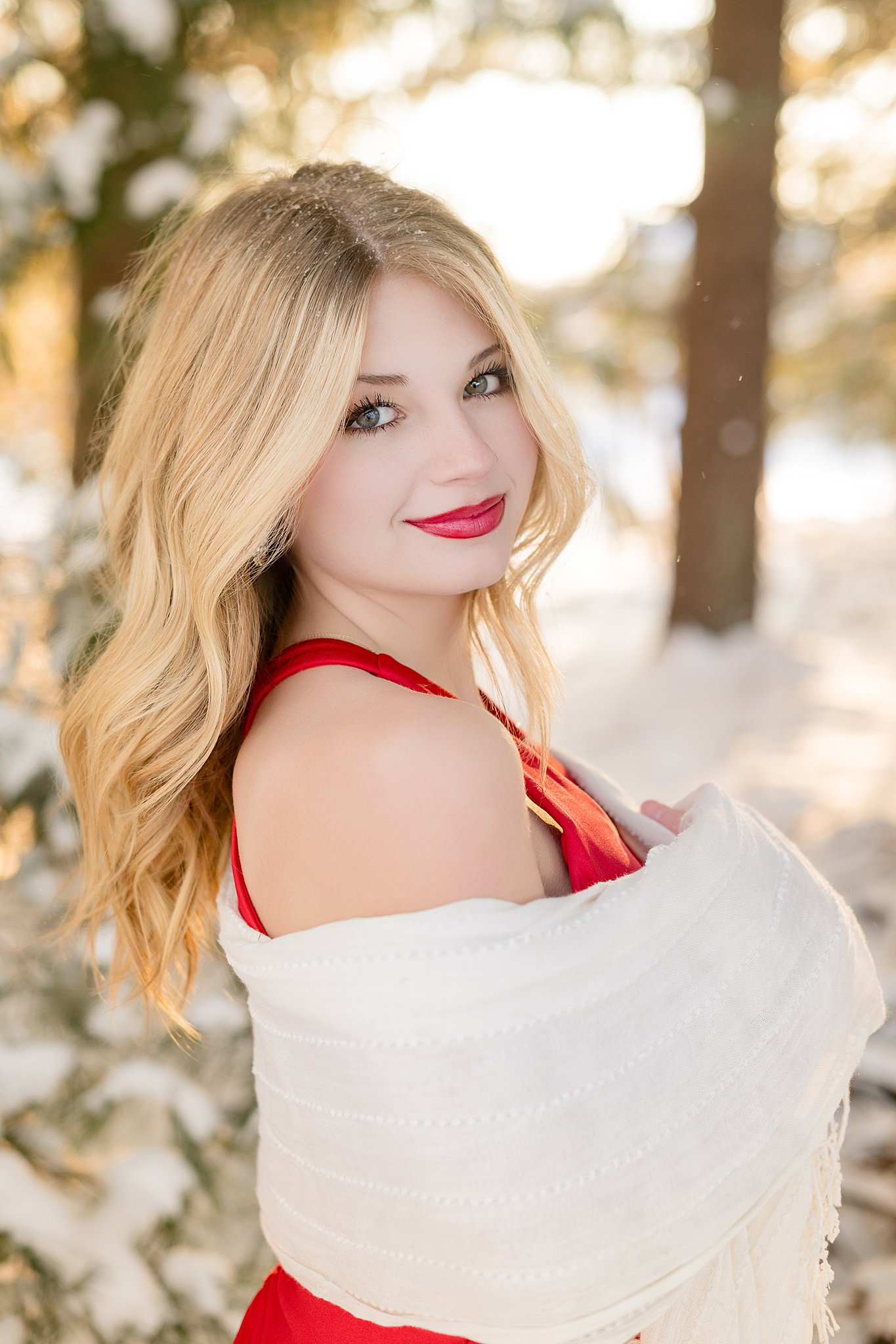 A high school senior stands in a red dress and wrapped in a white shawl in a snow covered forest smiling after visiting hair salons in Belpre, OH