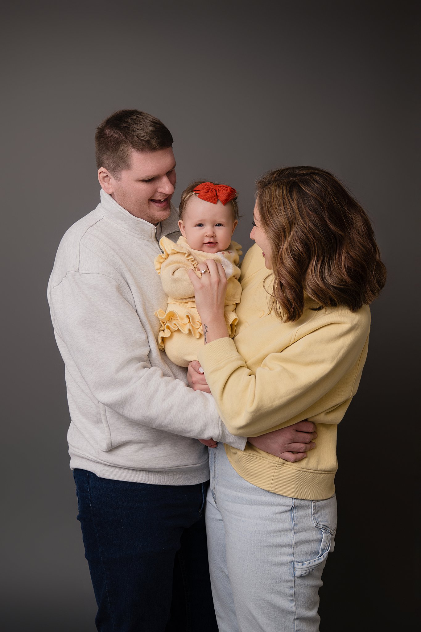Happy mom and dad in yellow and white stand in a studio playing with their infant daughter in a yellow dress after visiting kid-friendly restaurants in Belpre, OH