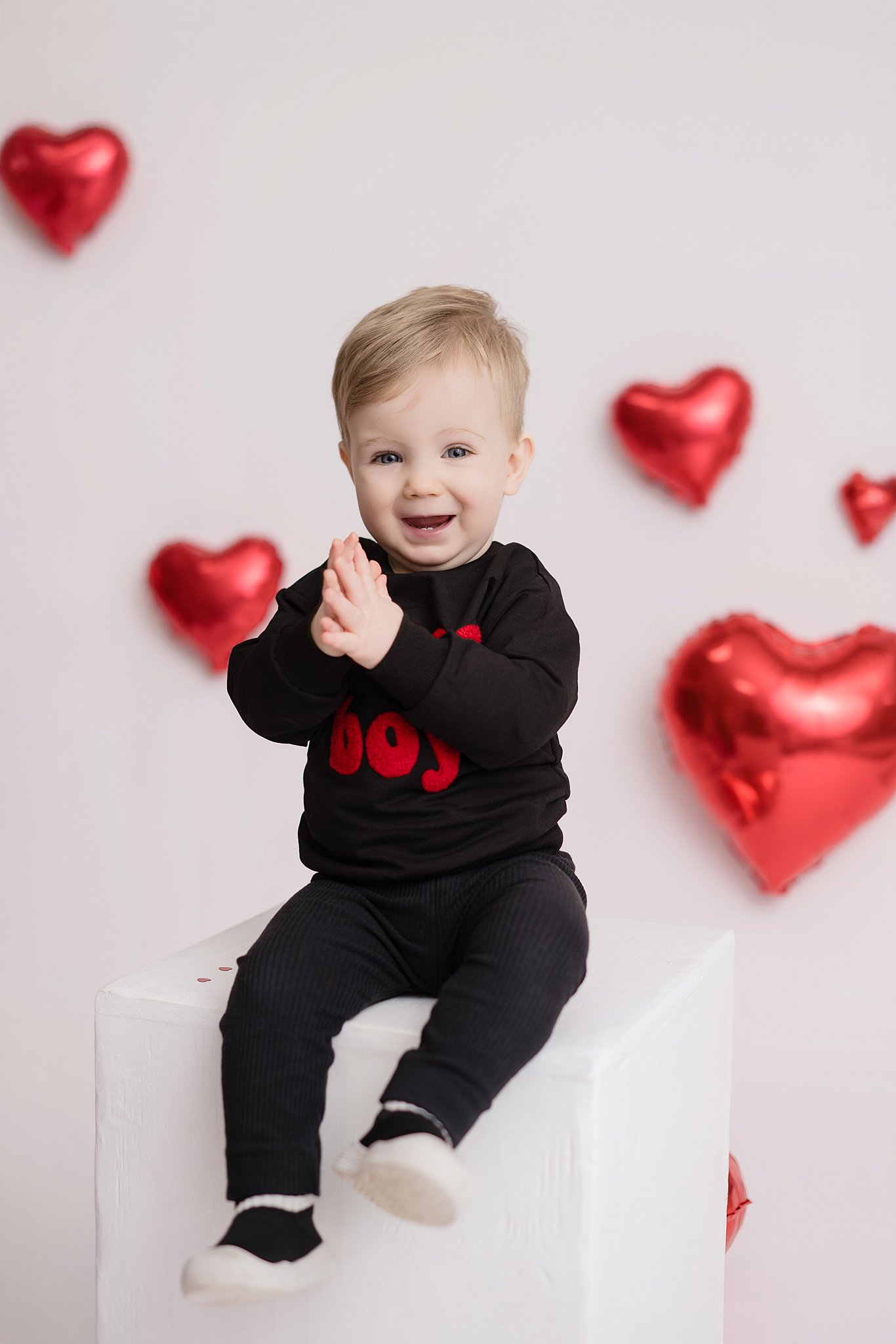 A happy toddler boy sits on a box in a studio clapping in a black outfit surrounded by balloon hearts after visiting a pediatric dentist in Belpre, Ohio