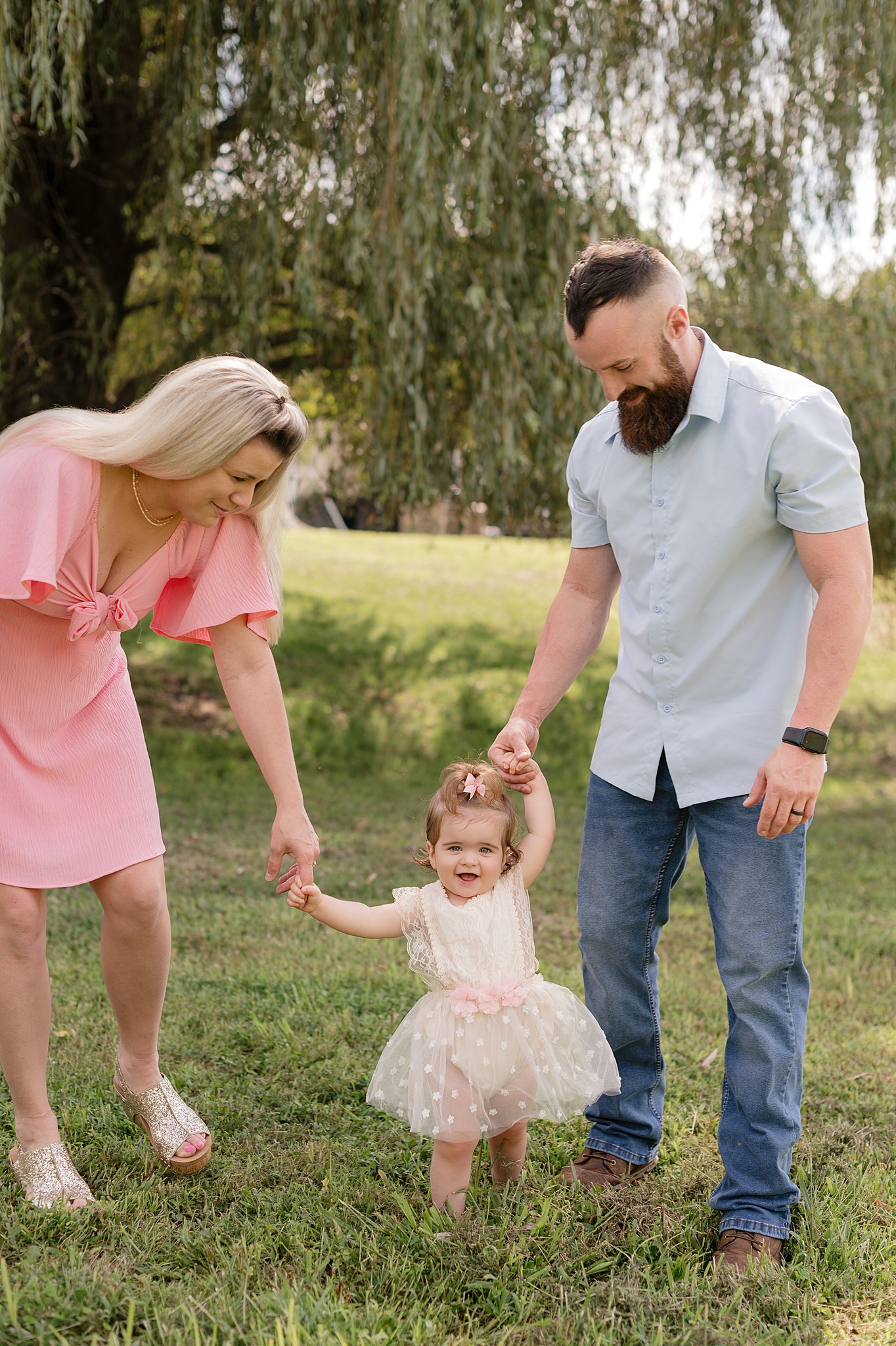 A happy couple walk their toddler daughter through a park in her white dress