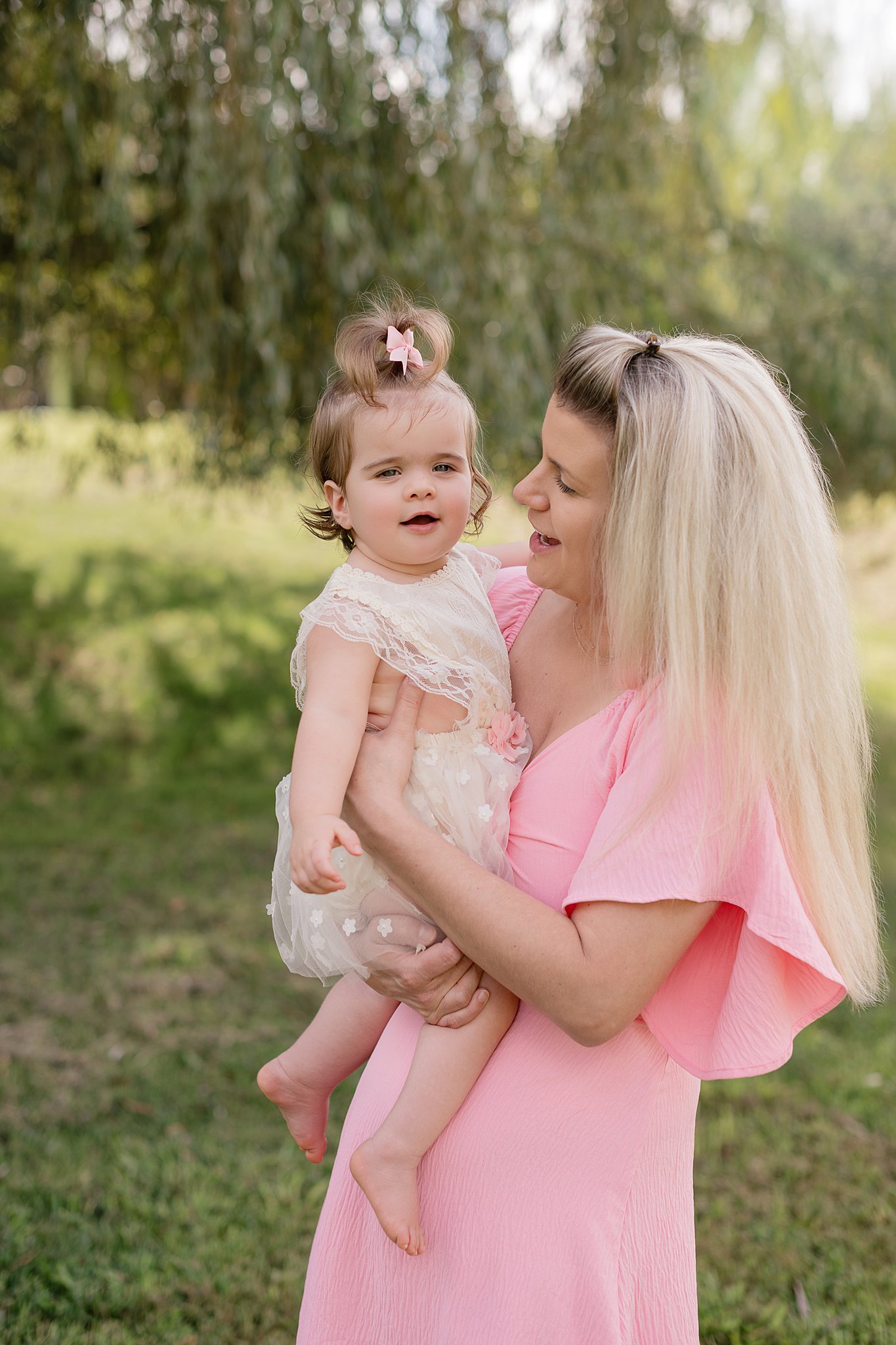 A happy mom in pink plays with her toddler daughter in a white dress in a park under a willow tree before heading to playgrounds in parkersburg wv