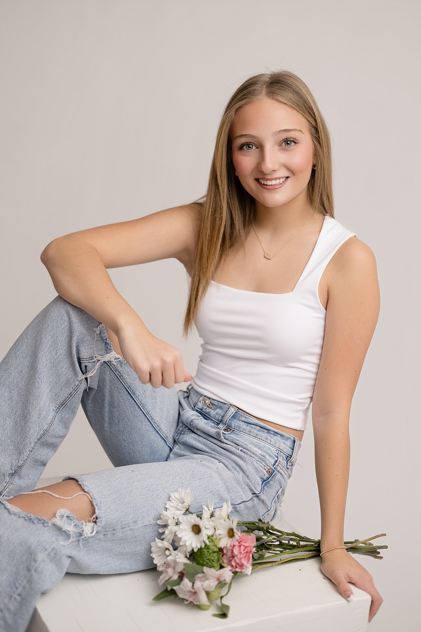 A happy high school senior sits on a white block in a studio in a white shirt and jeans after attending private schools in Parkersburg, WV