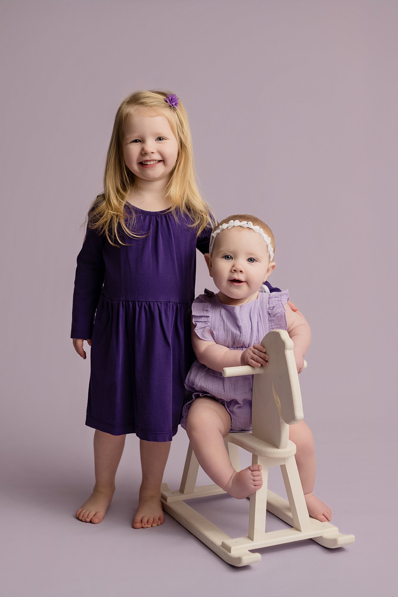 Toddler sisters in purple dresses in a studio with one on a rocking horse before some things to do with toddlers in Parkersburg, WV