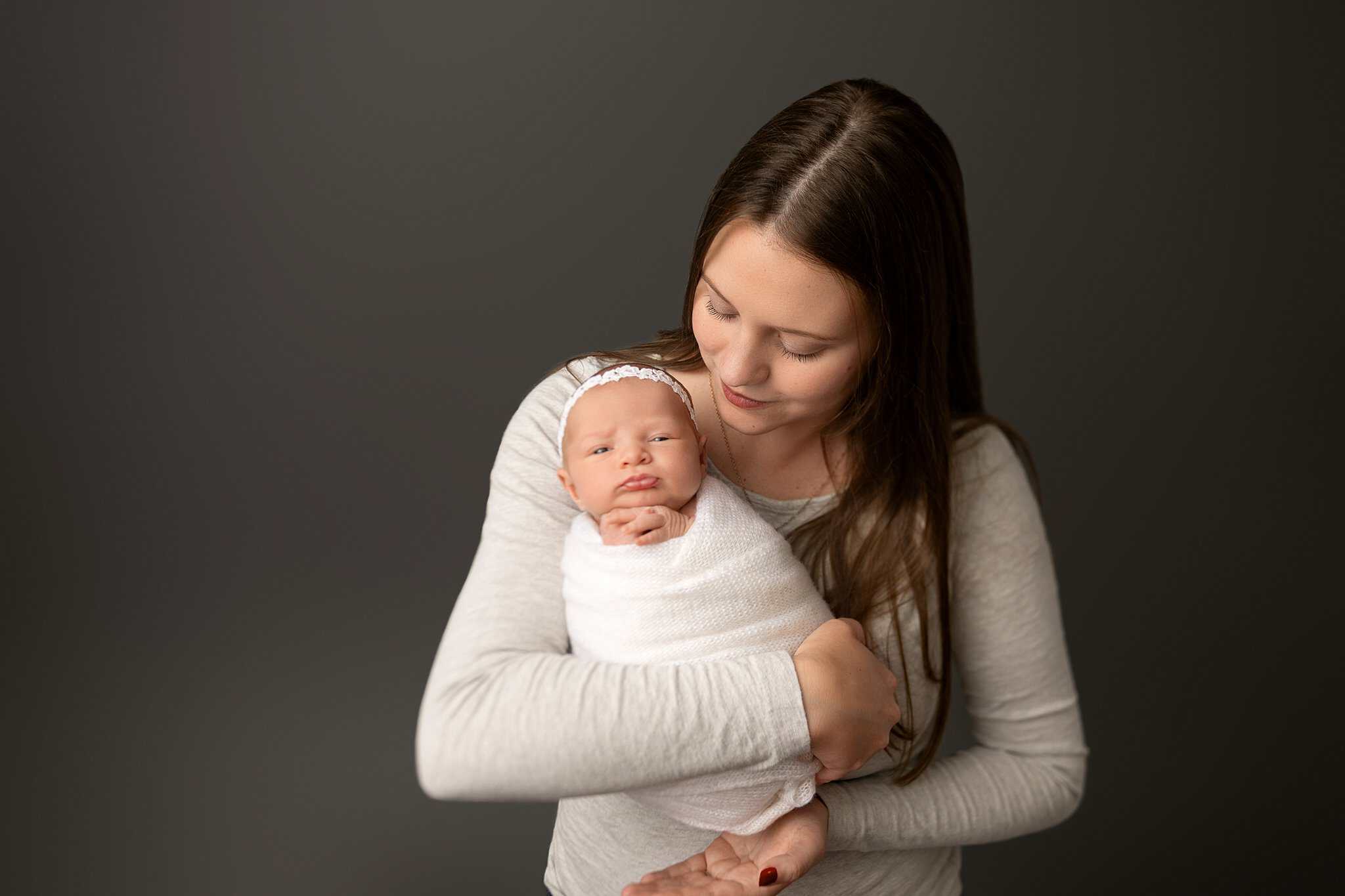A newborn baby in a white swaddle is held by mom in a grey shirt while standing in a studio