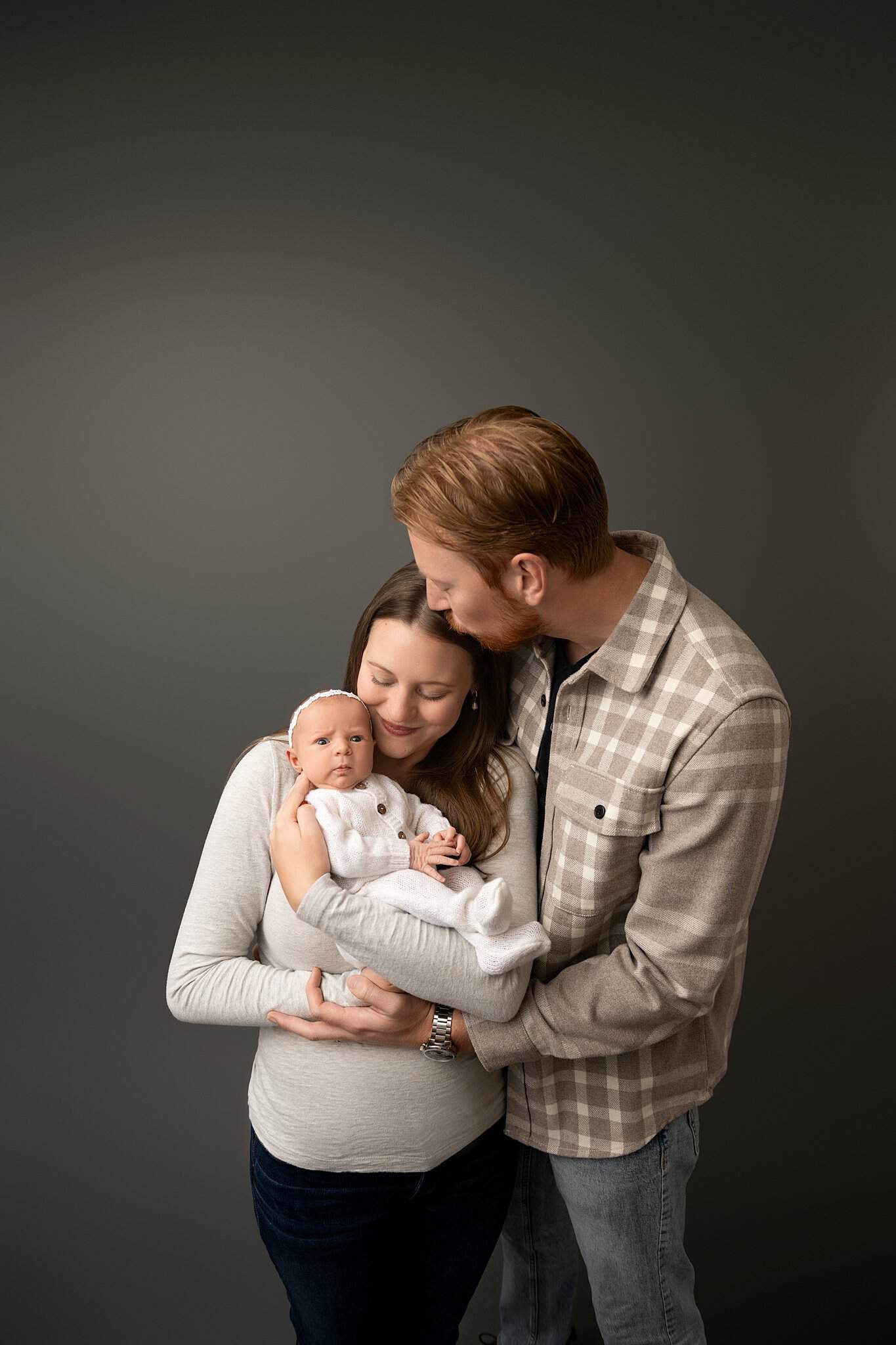 A happy new mom snuggles her awake newborn against her cheek while standing snuggling with her husband in plaid kissing her head after finding postpartum support in parkersburg, wv