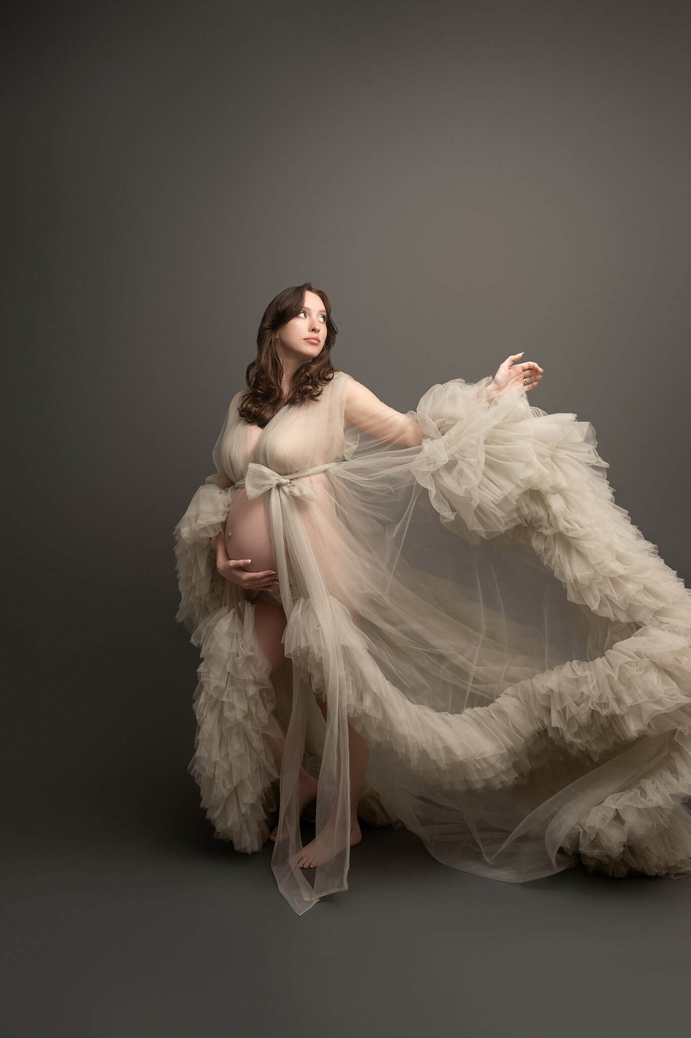 A pregnant woman plays with her long tule train with a hand under her bump in a studio
